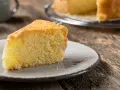 Una fetta di torta sabbiosa, soffice e dorata, su un piatto grigio. Sullo sfondo, un'altra torta intera su una gratella.