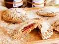 Biscotti integrali con marmellata, spolverati di zucchero a velo, su un tagliere di legno. Un morso rivela il ripieno rosso e invitante.