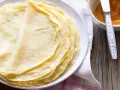 Impasto crepe dorate e sottili, pronte per essere farcite. Accanto, una ciotolina con marmellata e un coltellino.