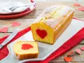 Plumcake con cuore rosso al centro, cosparso di zucchero a velo. Una fetta tagliata rivela la sorpresa. Decorazioni a forma di cuore completano l'atmosfera romantica.