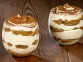 Due bicchieri di tiramisù senza uova benedetta fatto in casa, stratificato con crema e savoiardi, spolverato di cacao.