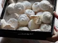 Scatola piena di soffici dolcetti al cocco, spolverati di zucchero a velo. Uno è spezzato, mostrando l'interno friabile.