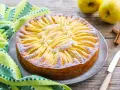 Torta di mele con yogurt, decorata con fettine di mela e zucchero a velo, su un piatto grigio, accanto a un tovagliolo a righe verdi e bianche.