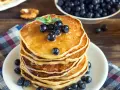 Pila di pancake vegani dorati, guarniti con mirtilli freschi e sciroppo d'acero, su un piatto bianco, con una ciotola di mirtilli e una bottiglia di sciroppo sullo sfondo.