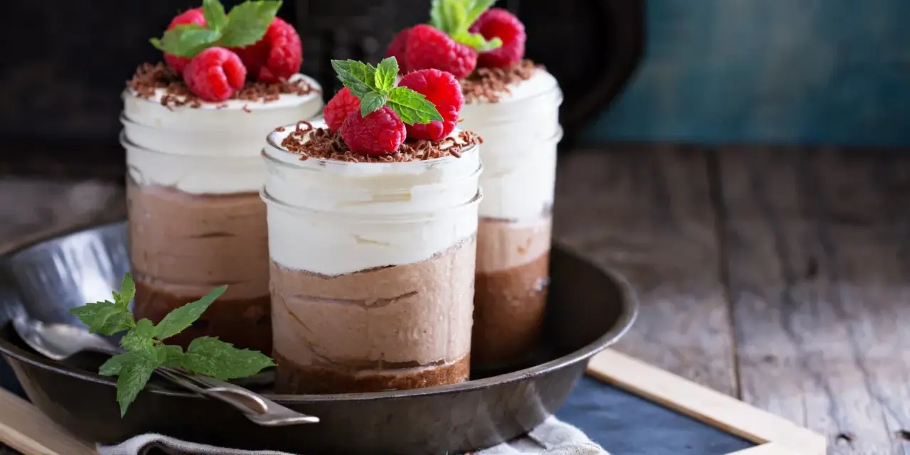 Tre deliziosi bicchierini di dolce freddo al cioccolato e panna, guarniti con lamponi freschi e scaglie di cioccolato.