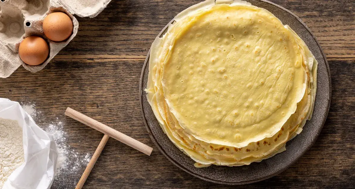 Pila di crepes dorate, pronte per essere farcite. Accanto, uova fresche e farina, ingredienti perfetti per crepes 2 uova.