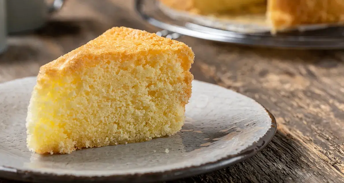 Una fetta di torta sabbiosa, soffice e dorata, su un piatto grigio. Sullo sfondo, un'altra torta intera su una gratella.