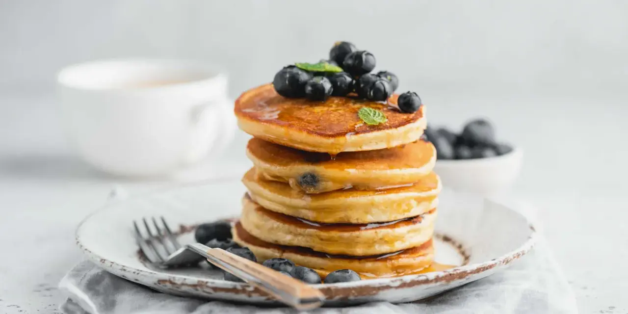 Pancake con farina di riso, guarniti con mirtilli freschi e sciroppo d'acero, su un piatto rustico con forchetta.
