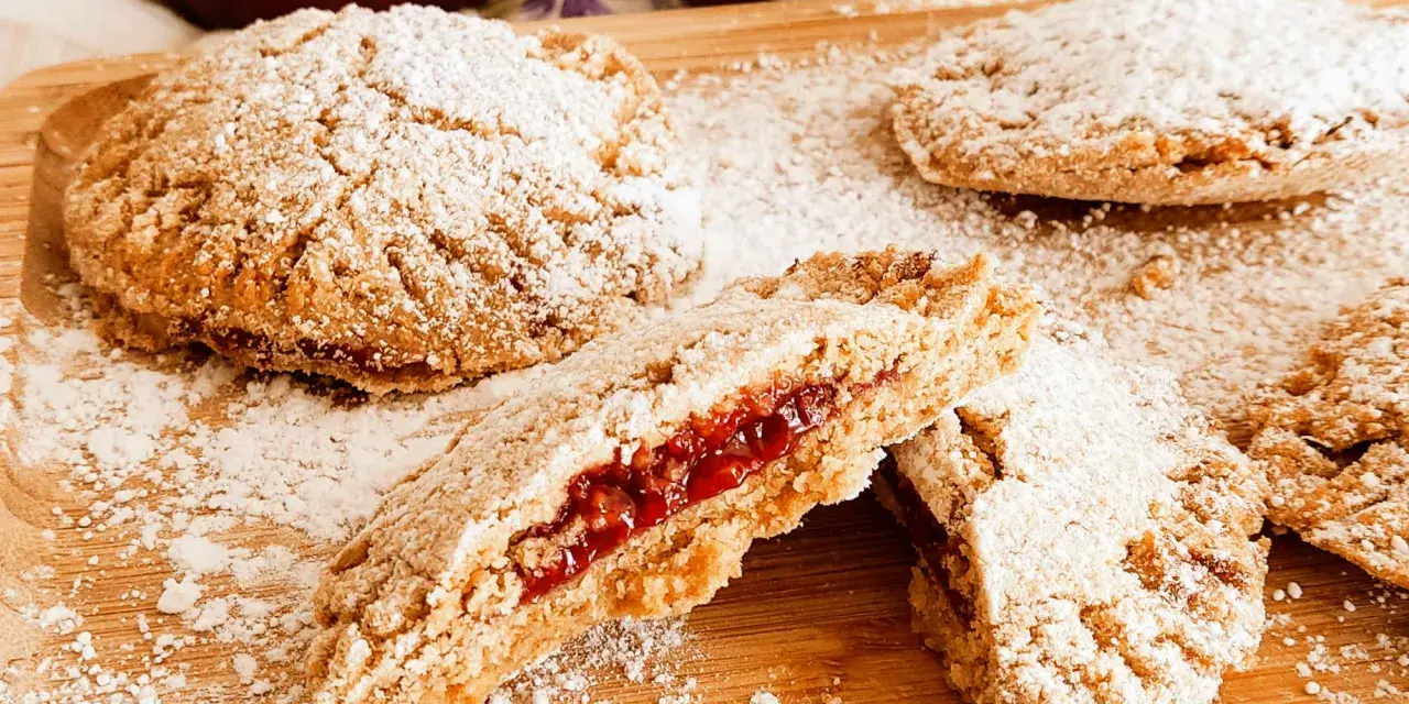 Biscotti integrali con marmellata, spolverati di zucchero a velo, su un tagliere di legno. Un morso rivela il ripieno rosso e invitante.
