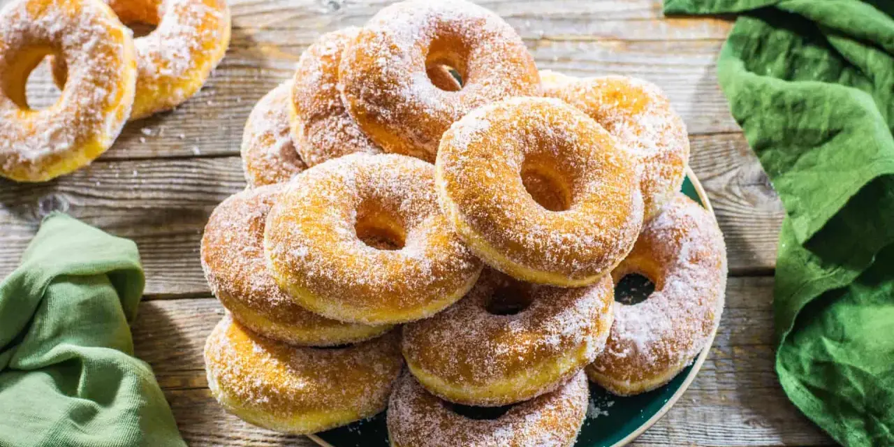 Un piatto pieno di graffe napoletane, dolci soffici e dorati, cosparse di zucchero a velo, su un tavolo di legno rustico.