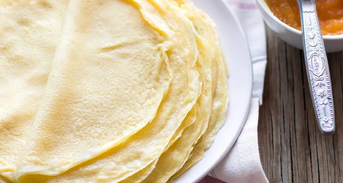 Impasto crepe dorate e sottili, pronte per essere farcite. Accanto, una ciotolina con marmellata e un coltellino.