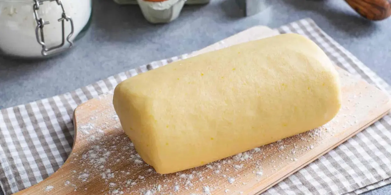 Rotolo di pasta frolla con strutto, pronto per essere trasformato in deliziosi biscotti. Ingredienti base visibili: farina, uova.
