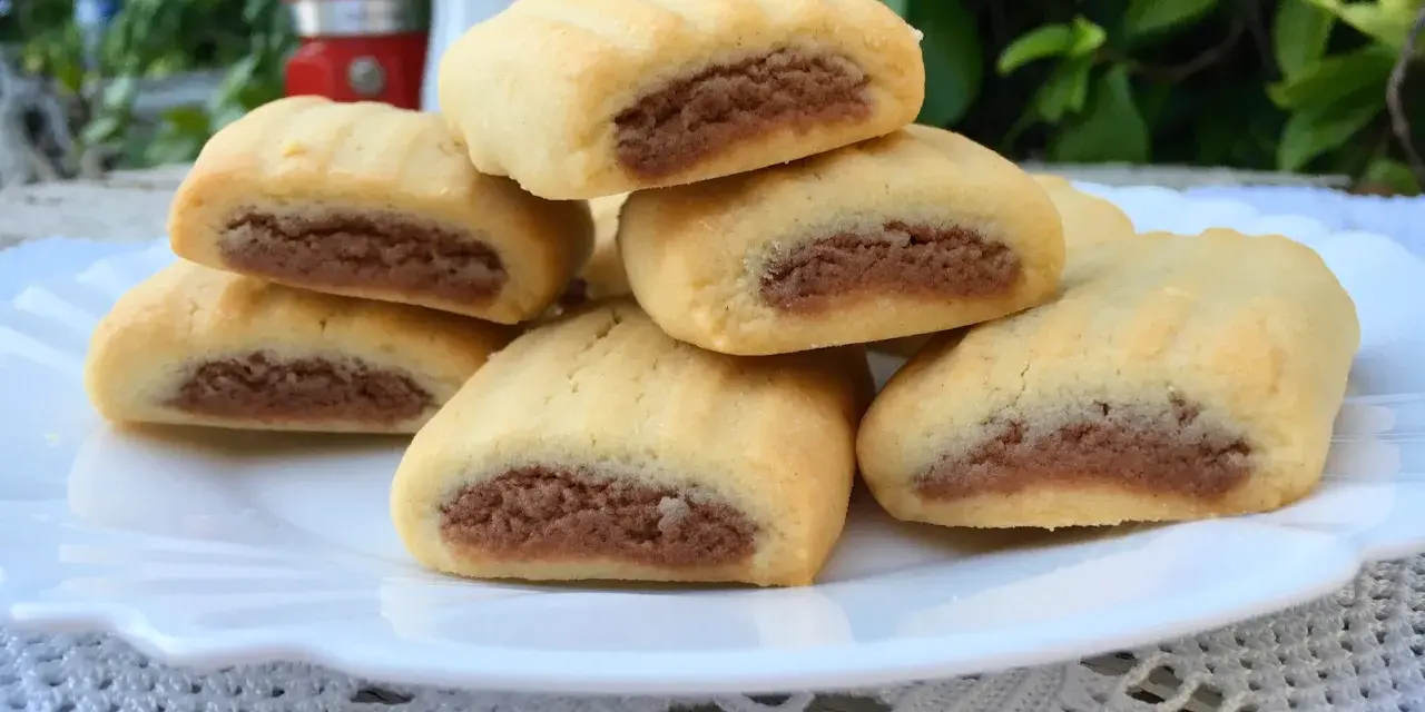 Biscotti nascondini impilati su un piatto bianco, con un ripieno scuro visibile. Sullo sfondo, una moka rossa e foglie verdi.