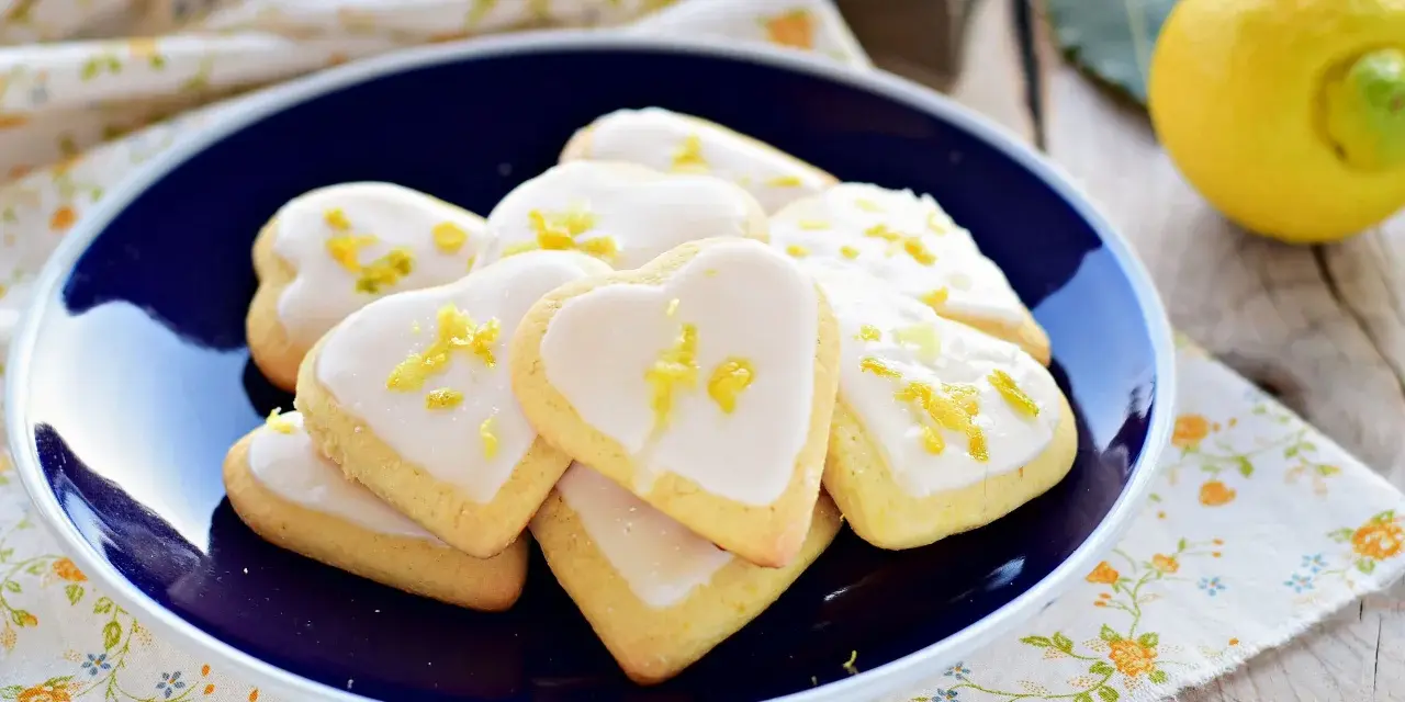 Cuore di biscotti al limone glassati, decorati con scorza di limone, su un piatto blu.