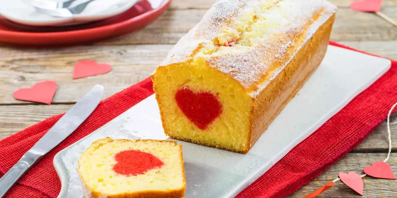 Plumcake con cuore rosso al centro, cosparso di zucchero a velo. Una fetta tagliata rivela la sorpresa. Decorazioni a forma di cuore completano l'atmosfera romantica.