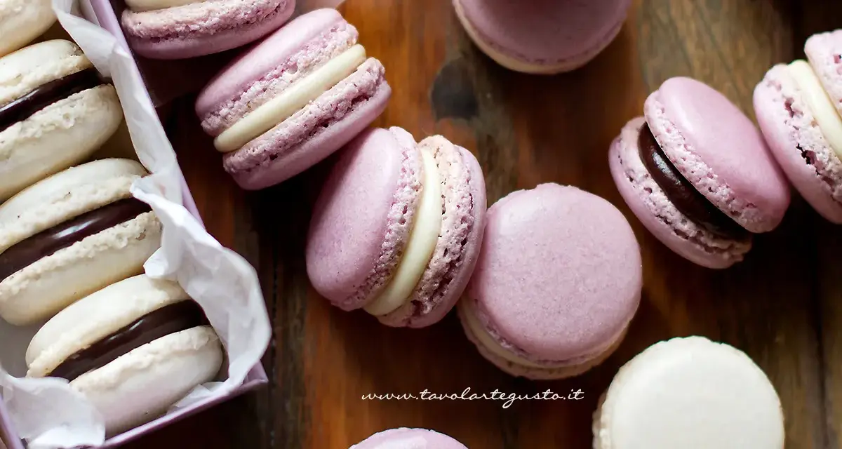 Deliziosi macaron dolci rosa e bianchi, alcuni con ripieno al cioccolato, sparsi su un tagliere di legno.
