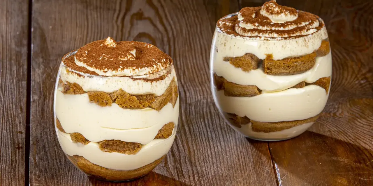 Due bicchieri di tiramisù senza uova benedetta fatto in casa, stratificato con crema e savoiardi, spolverato di cacao.