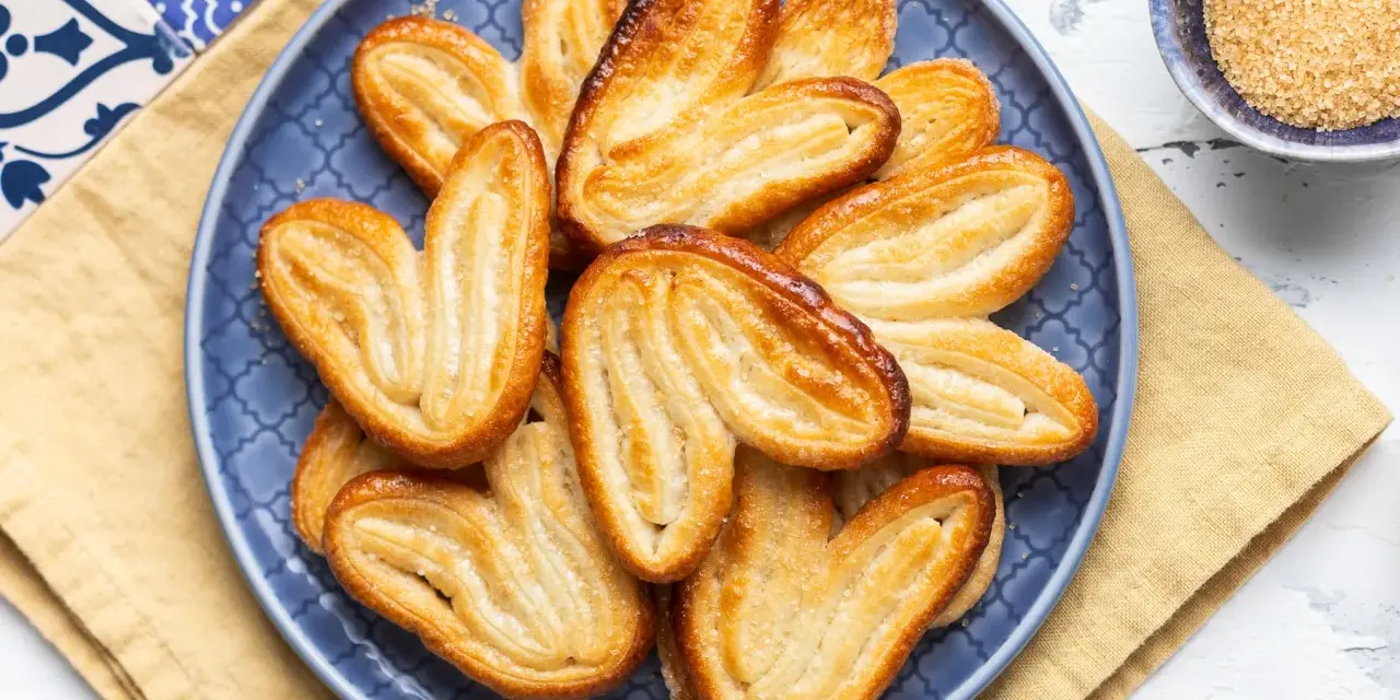 Un piatto blu pieno di croccanti ventagli di pasta sfoglia, dorati e zuccherati, pronti per essere gustati.
