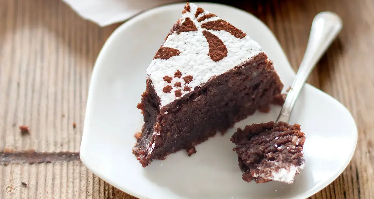 Una fetta di torta caprese ricetta facile, cosparsa di zucchero a velo e cacao, su un piatto bianco a forma di cuore.