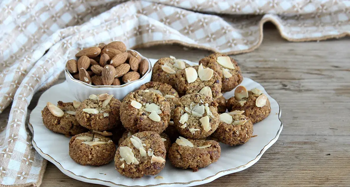 Biscotti senza zucchero, dorati e croccanti, guarniti con mandorle a lamelle. Accanto, una ciotola piena di mandorle intere.