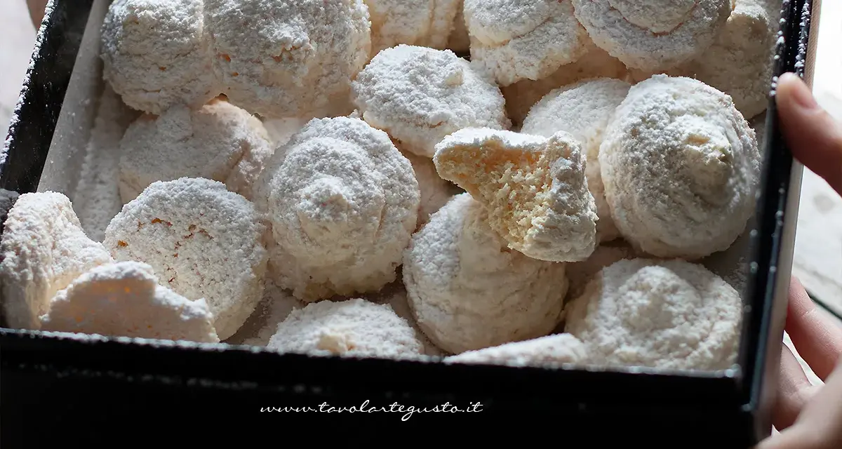 Scatola piena di soffici dolcetti al cocco, spolverati di zucchero a velo. Uno è spezzato, mostrando l'interno friabile.