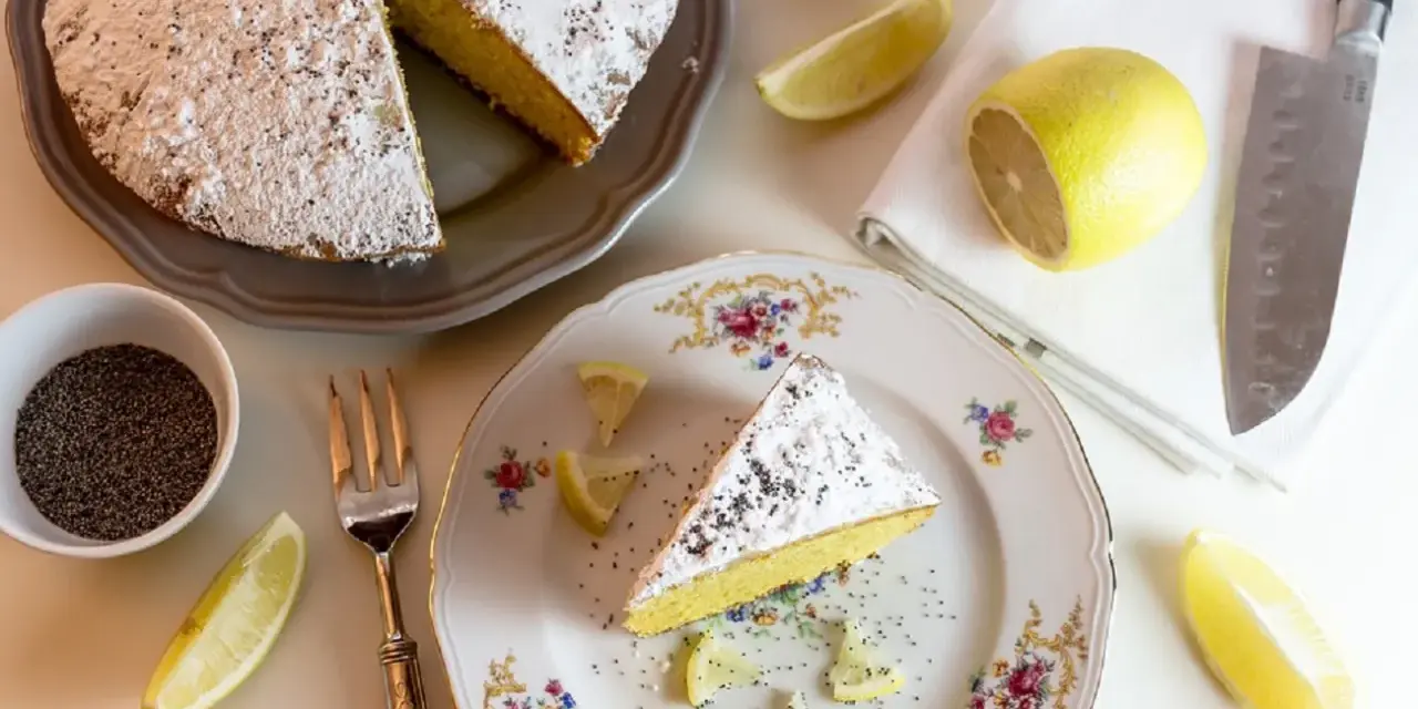Una fetta di torta al limone cosparsa di zucchero a velo e semi di papavero, guarnita con spicchi di limone fresco.