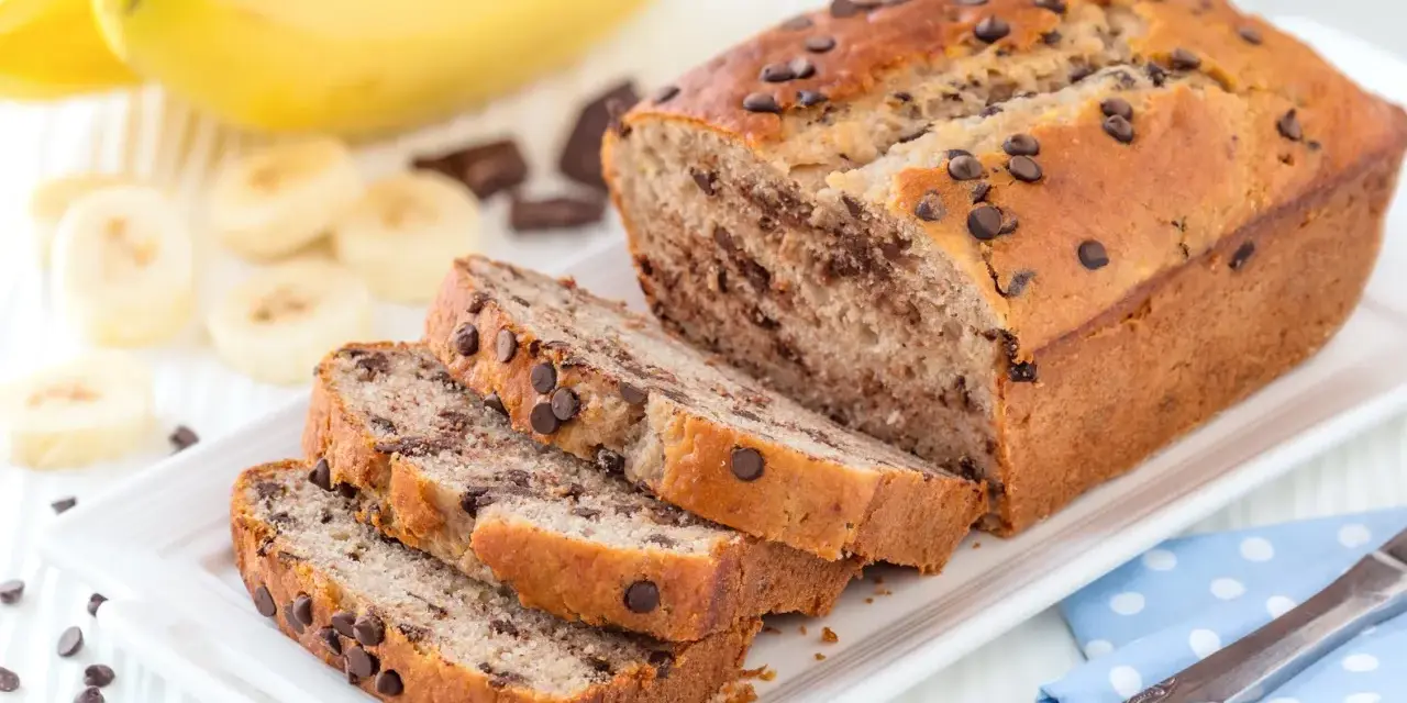 Fette di plumcake banana e cioccolato su un piatto bianco, con banane e gocce di cioccolato sullo sfondo.
