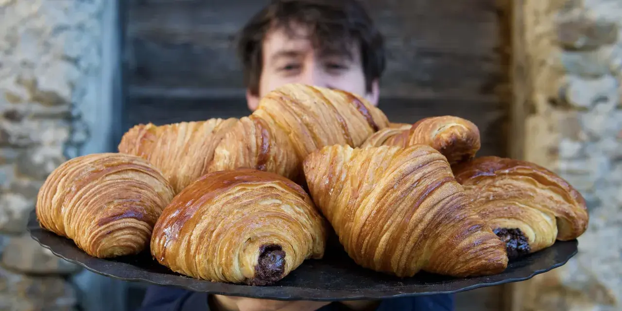 Un ragazzo sorride dietro un vassoio pieno di croissant brioche dorati e fragranti, alcuni con ripieno di cioccolato.