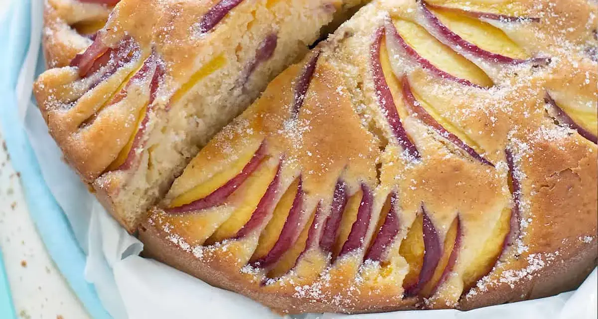 Una deliziosa torta di pesche e yogurt greco, con fette di pesca disposte a raggiera sulla superficie dorata e cosparsa di zucchero a velo.