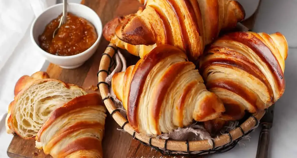 Croissant dorati e sfogliati, un invito a scoprire la loro origine. Marmellata in ciotola e una tazza bianca completano la scena.