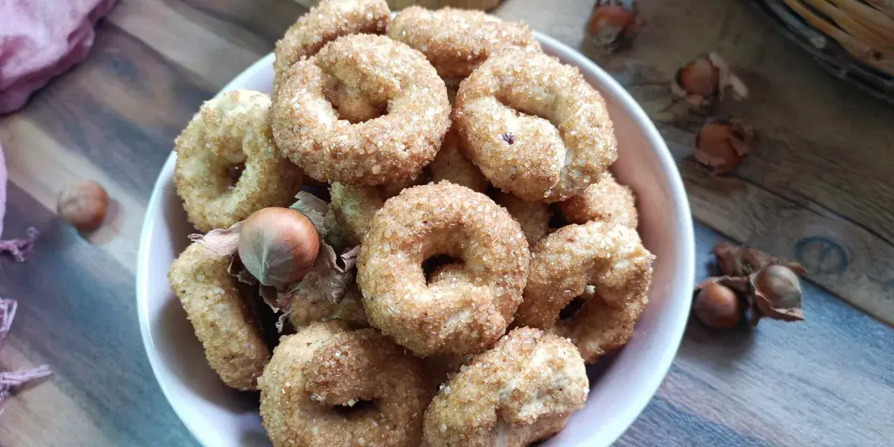 Ciambelline al vino cosparse di zucchero, con nocciole sparse sul tavolo di legno.
