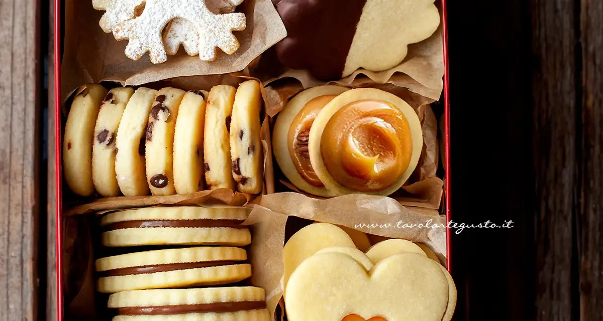 Scatola piena di biscotti al burro: a fiocco di neve, con gocce di cioccolato, ripieni di caramello, a forma di cuore e ricoperti di cioccolato.