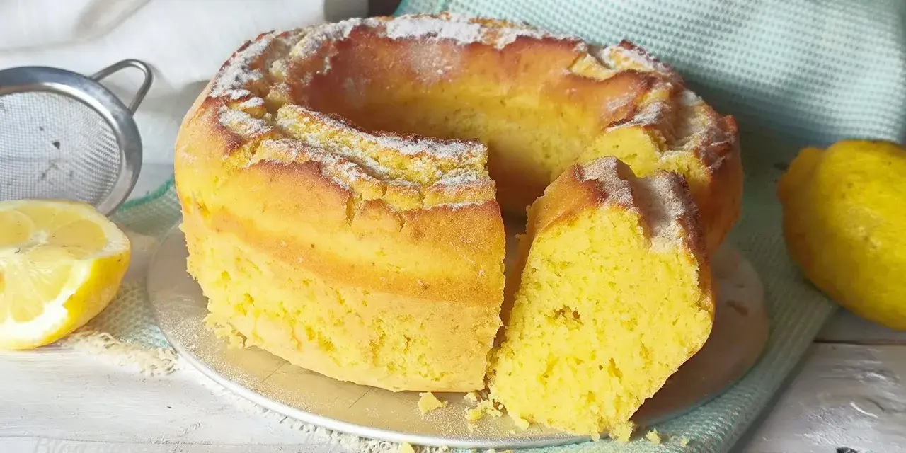Torta soffice e dorata, spolverata di zucchero a velo, preparata con dolci con eritritolo. Accanto, limoni freschi e un setaccio.