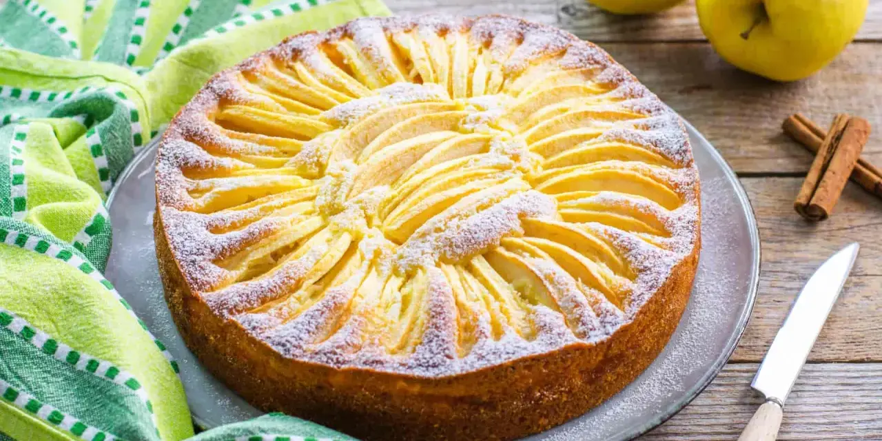 Torta di mele con yogurt, decorata con fettine di mela e zucchero a velo, su un piatto grigio, accanto a un tovagliolo a righe verdi e bianche.