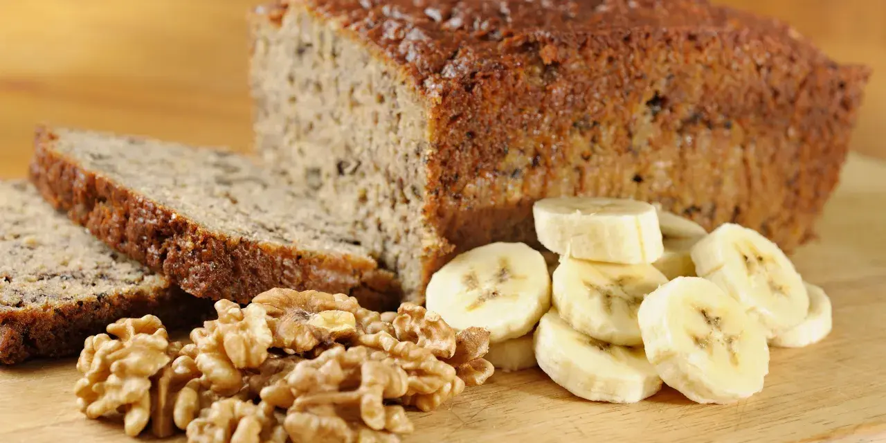 Fetta di banana bread senza uova, con noci e banane a fette, su tagliere di legno.