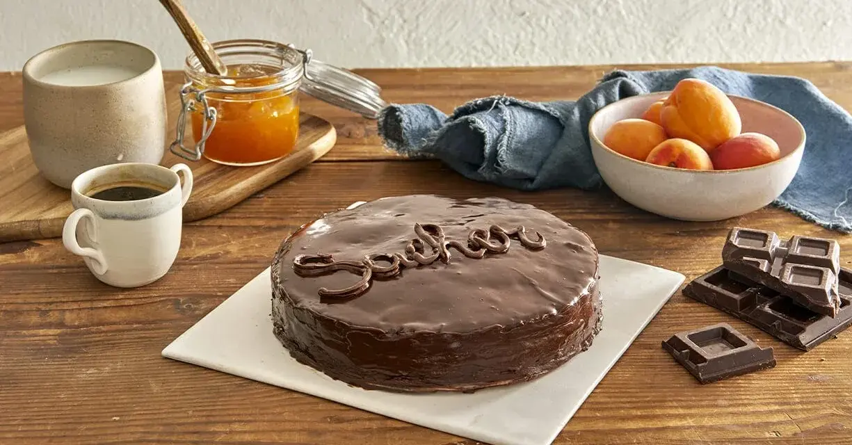 Torta Sacher, un classico viennese, con la sua glassa lucida e il nome scritto in cioccolato. Accanto, albicocche, marmellata e caffè, ingredienti che evocano le sue origini.
