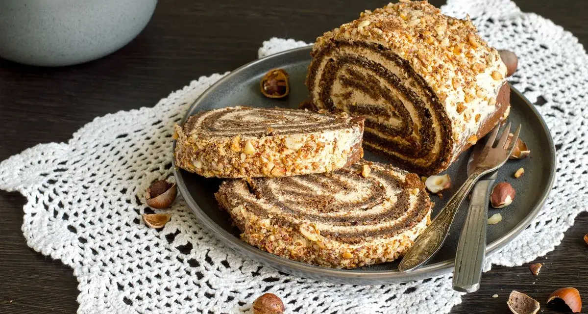 Rotolo alla Nutella ricoperto di granella di nocciole, servito su un piatto con due fette tagliate.