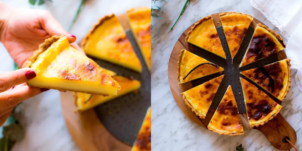 Una mano prende una fetta di flan parisien dorato. La torta intera, tagliata a spicchi, è su un tagliere di legno, decorata con un fiore e un rametto di eucalipto.