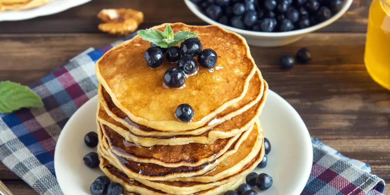 Pila di pancake vegani dorati, guarniti con mirtilli freschi e sciroppo d'acero, su un piatto bianco, con una ciotola di mirtilli e una bottiglia di sciroppo sullo sfondo.