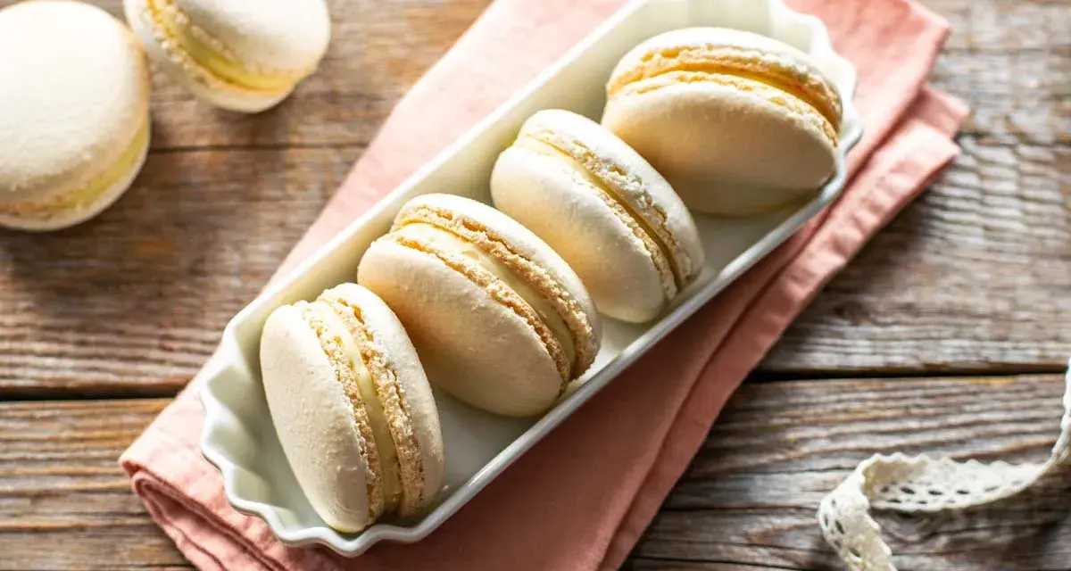 Macarons bianchi e dorati, perfetti esempi di come sono fatti i macarons, con ripieno cremoso, su un piatto bianco e tovagliolo rosa.