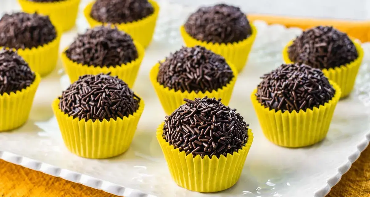 Deliziosi dolcetti brasiliani, brigadeiros al cioccolato, ricoperti di codette, serviti in pirottini gialli su un piatto bianco.