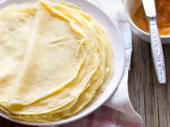 Impasto crepe dorate e sottili, pronte per essere farcite. Accanto, una ciotolina con marmellata e un coltellino.