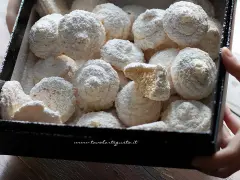 Scatola piena di soffici dolcetti al cocco, spolverati di zucchero a velo. Uno è spezzato, mostrando l'interno friabile.