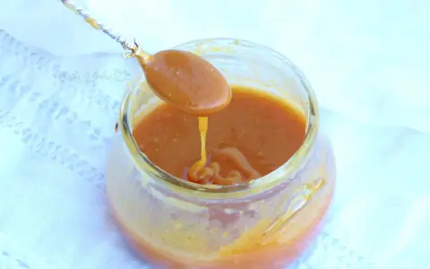 Un cucchiaio raccoglie un denso caramello salato da un barattolo di vetro. La salsa dorata cola lentamente, invitando a un assaggio goloso.