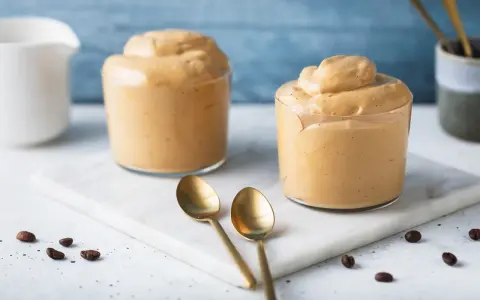 Due bicchieri di soffice crema di caffè, perfetta ricetta crema di caffè senza panna e senza caffè solubile. Due cucchiaini dorati e chicchi di caffè completano la scena.