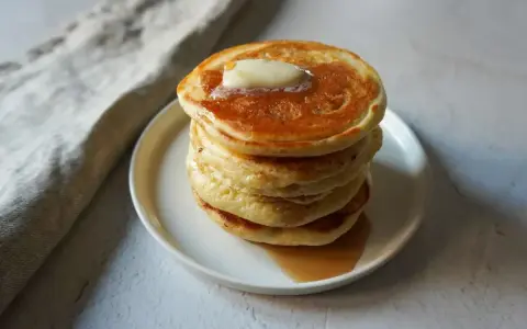 Pancakes dorati e soffici, impilati su un piatto bianco, con una noce di burro che si scioglie in cima e sciroppo d'acero che cola. Sembrano pronti per essere gustati, un vero piacere per gli occhi e per il palato, come un pancake in bottiglia pronto d...