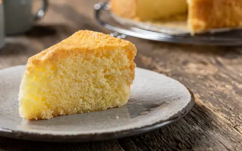 Una fetta di torta sabbiosa, soffice e dorata, su un piatto grigio. Sullo sfondo, un'altra torta intera su una gratella.