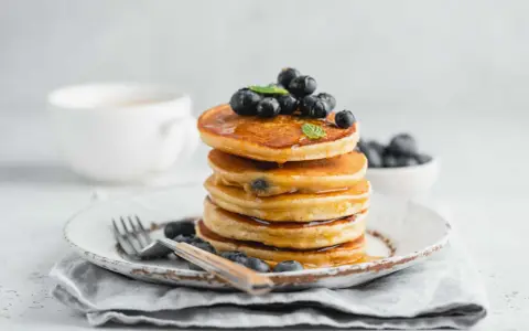 Pancake con farina di riso, guarniti con mirtilli freschi e sciroppo d'acero, su un piatto rustico con forchetta.