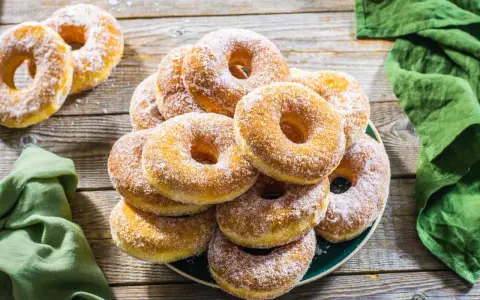 Un piatto pieno di graffe napoletane, dolci soffici e dorati, cosparse di zucchero a velo, su un tavolo di legno rustico.