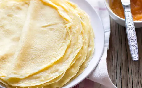 Impasto crepe dorate e sottili, pronte per essere farcite. Accanto, una ciotolina con marmellata e un coltellino.